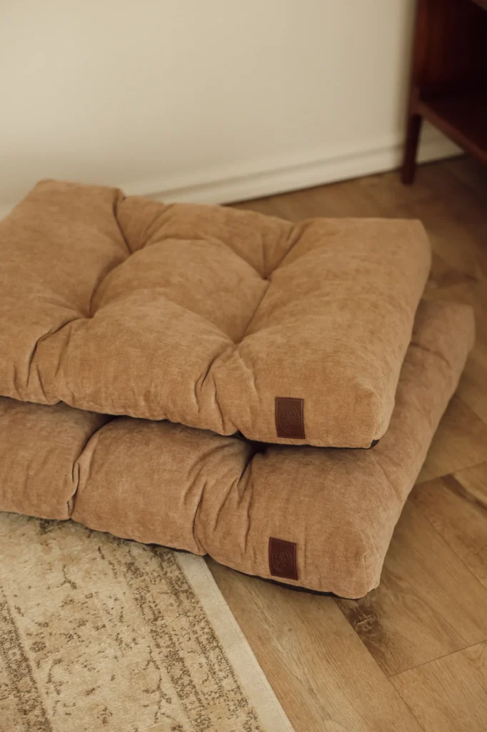 Two beige Atelier dog beds stacked on a wooden floor in a living room