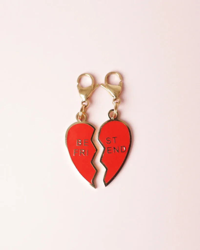 Pareja de charms Best Friends de corazón partido rojo con mosquetones dorados sobre fondo rosa palo