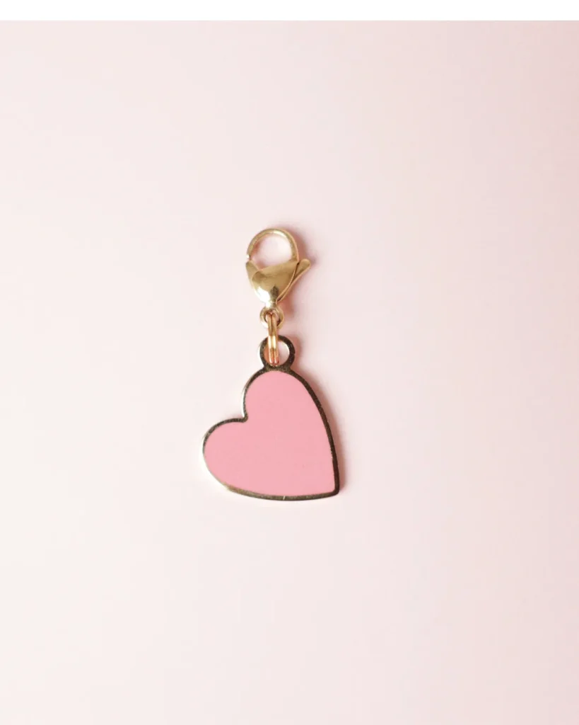 Charm de corazón rosa esmaltado con mosquetón dorado sobre fondo rosa palo