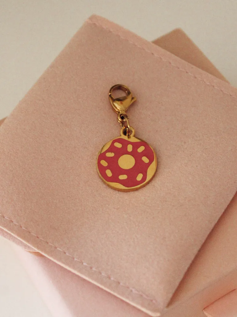 Charm de dónut con glaseado rosa y virutas y mosquetón dorado sobre una funda rosa palo