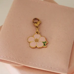 Charm de flor Bloom rosa palo de cinco pétalos con hojas verdes y mosquetón dorado sobre una funda rosa palo