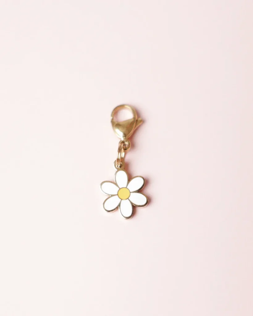 Charm de flor margarita blanca con el centro amarillo y mosquetón dorado sobre fondo rosa palo