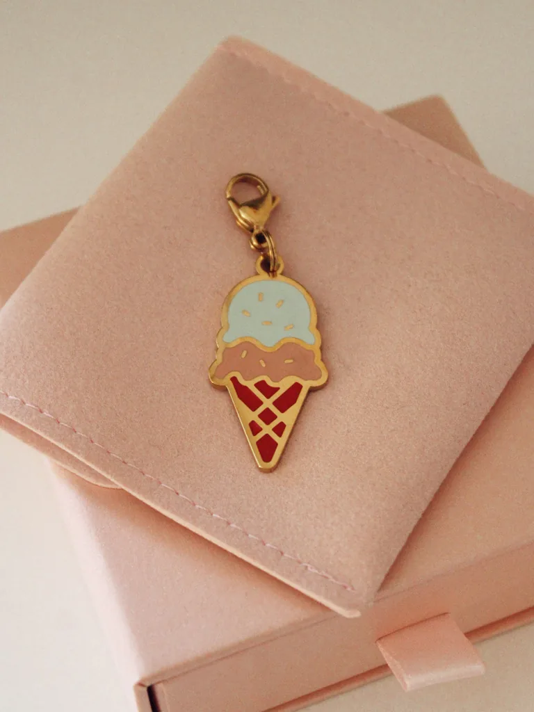 Charm de cucurucho de helado de dos bolas, azul y rosa, con mosquetón dorado sobre una funda rosa palo