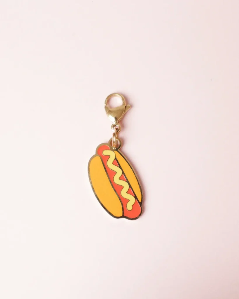 Charm de perrito caliente con mostaza amarilla y mosquetón dorado sobre fondo rosa palo