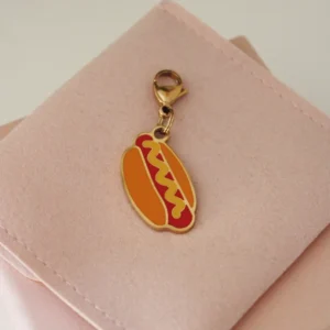 Charm de perrito caliente con mostaza amarilla y mosquetón dorado sobre una funda rosa palo