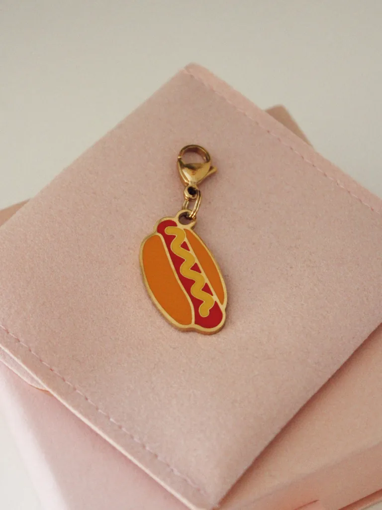Charm de perrito caliente con mostaza amarilla y mosquetón dorado sobre una funda rosa palo
