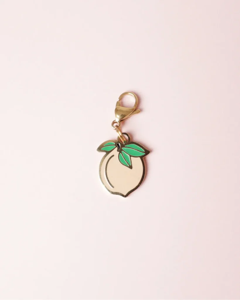 Charm de melocotón rosa palo con hojas verdes y mosquetón dorado sobre fondo rosa palo