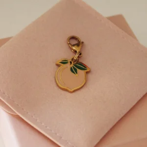 Charm de melocotón rosa palo con hojas verdes y mosquetón dorado sobre una funda rosa palo