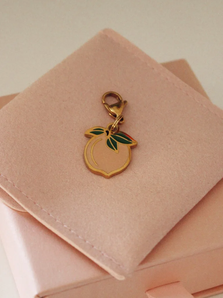 Charm de melocotón rosa palo con hojas verdes y mosquetón dorado sobre una funda rosa palo