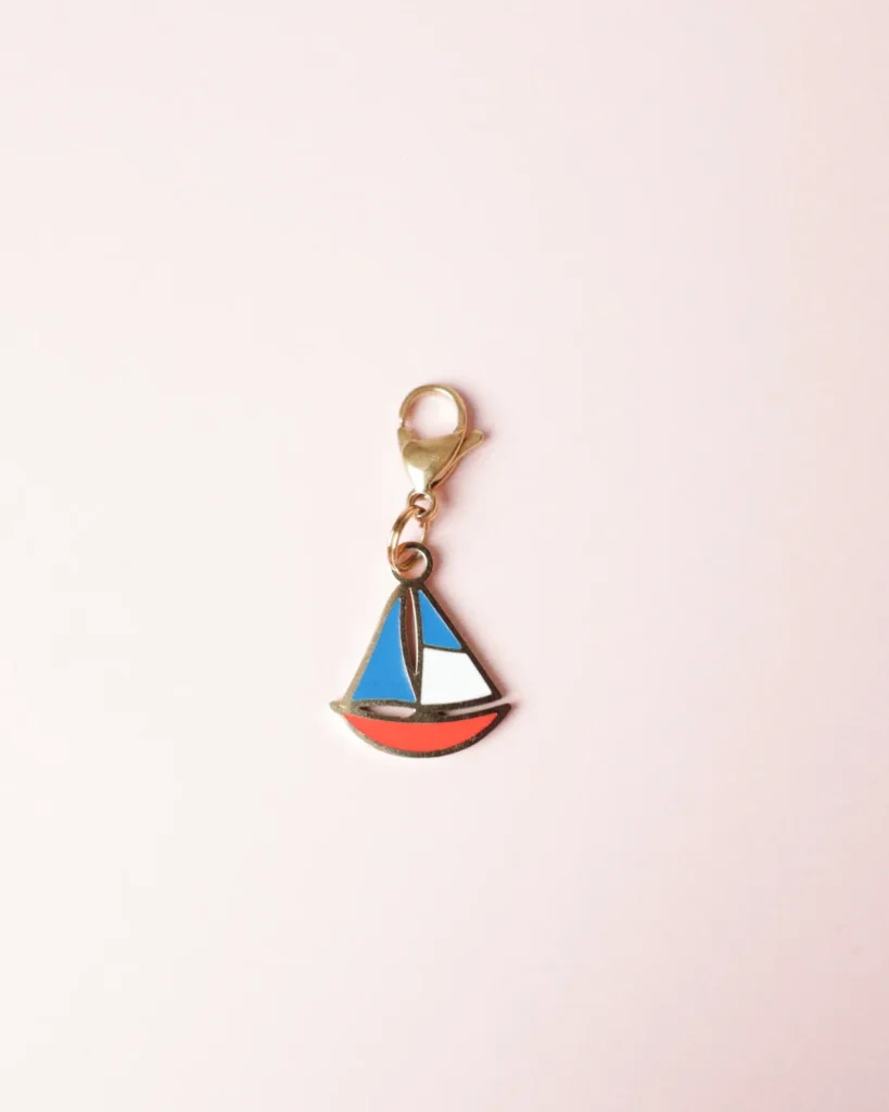 Charm de velero rojo, blanco y azul con mosquetón dorado sobre fondo rosa palo