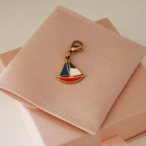 Charm de velero rojo, blanco y azul con mosquetón dorado sobre una funda rosa palo