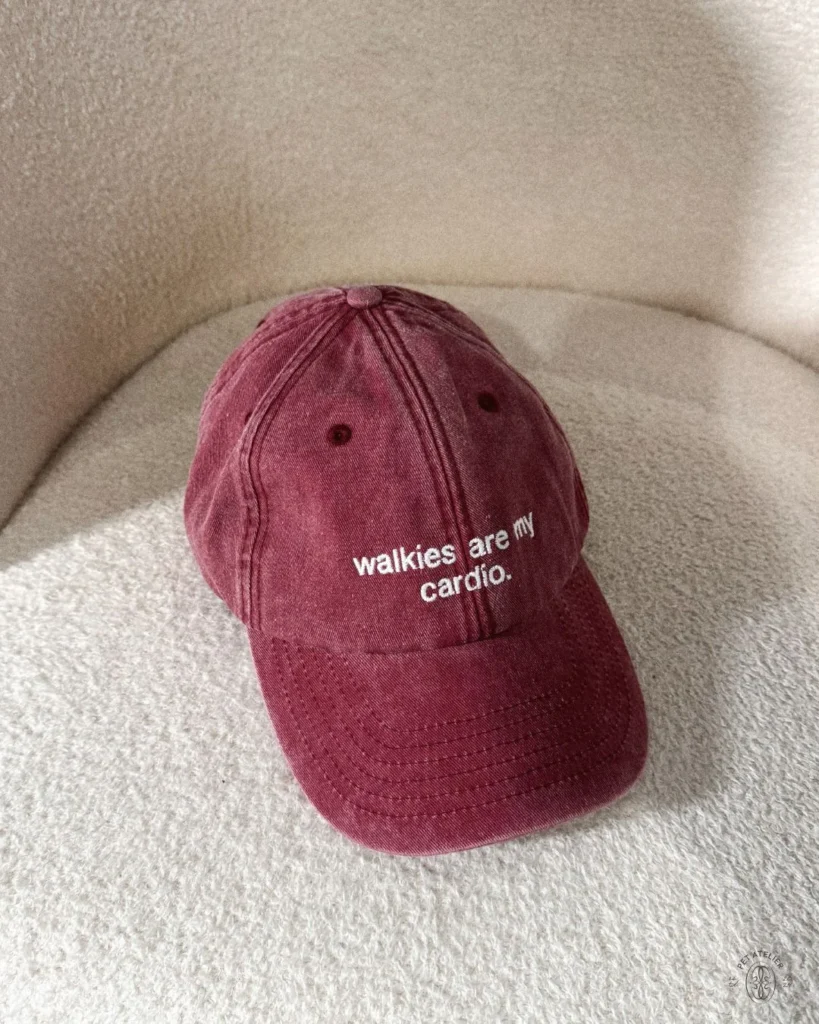 Washed red cap with “walkies are my cardio.” embroidered in white, on a cream bouclé chair