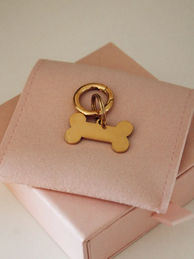 Placa identificativa dorada pequeña para perro en forma de hueso, personalizada y grabada con láser, con anilla dorada sobre una funda rosa palo