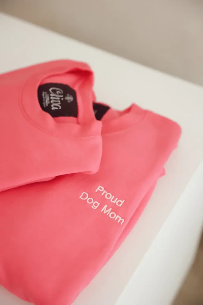 Dos sudaderas Dog Mom rosas dobladas, con «Proud Dog Mom» bordado en blanco y la etiqueta Oliva en el cuello